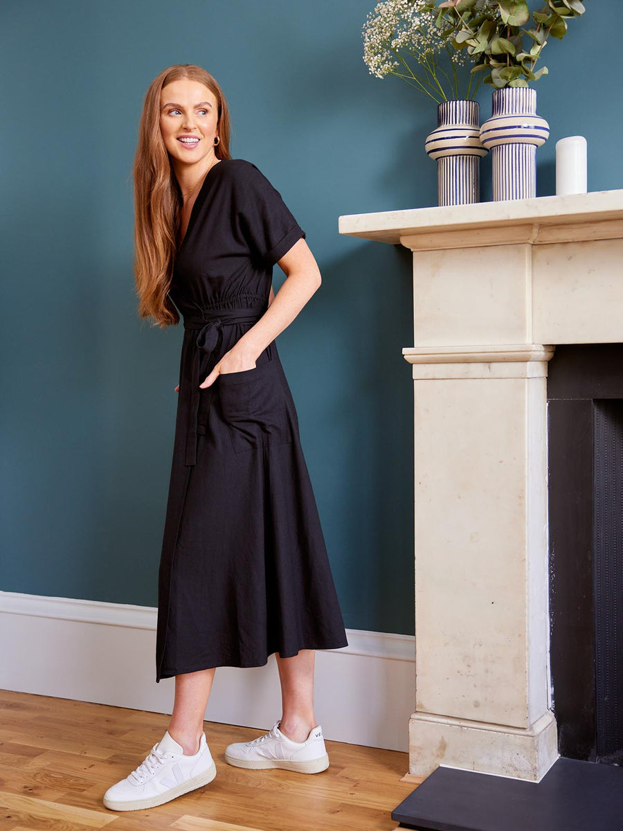 A model stands in a living area next to a large fireplace with plants on the mantlepiece. They have their hands in the pockets of the Daisy wrap dress that they are wearing.