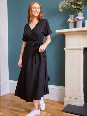 A model stands in a living area next to a large fireplace with plants on the mantlepiece. They are wearing the sustainable Daisy wrap dress from Unfolded. 