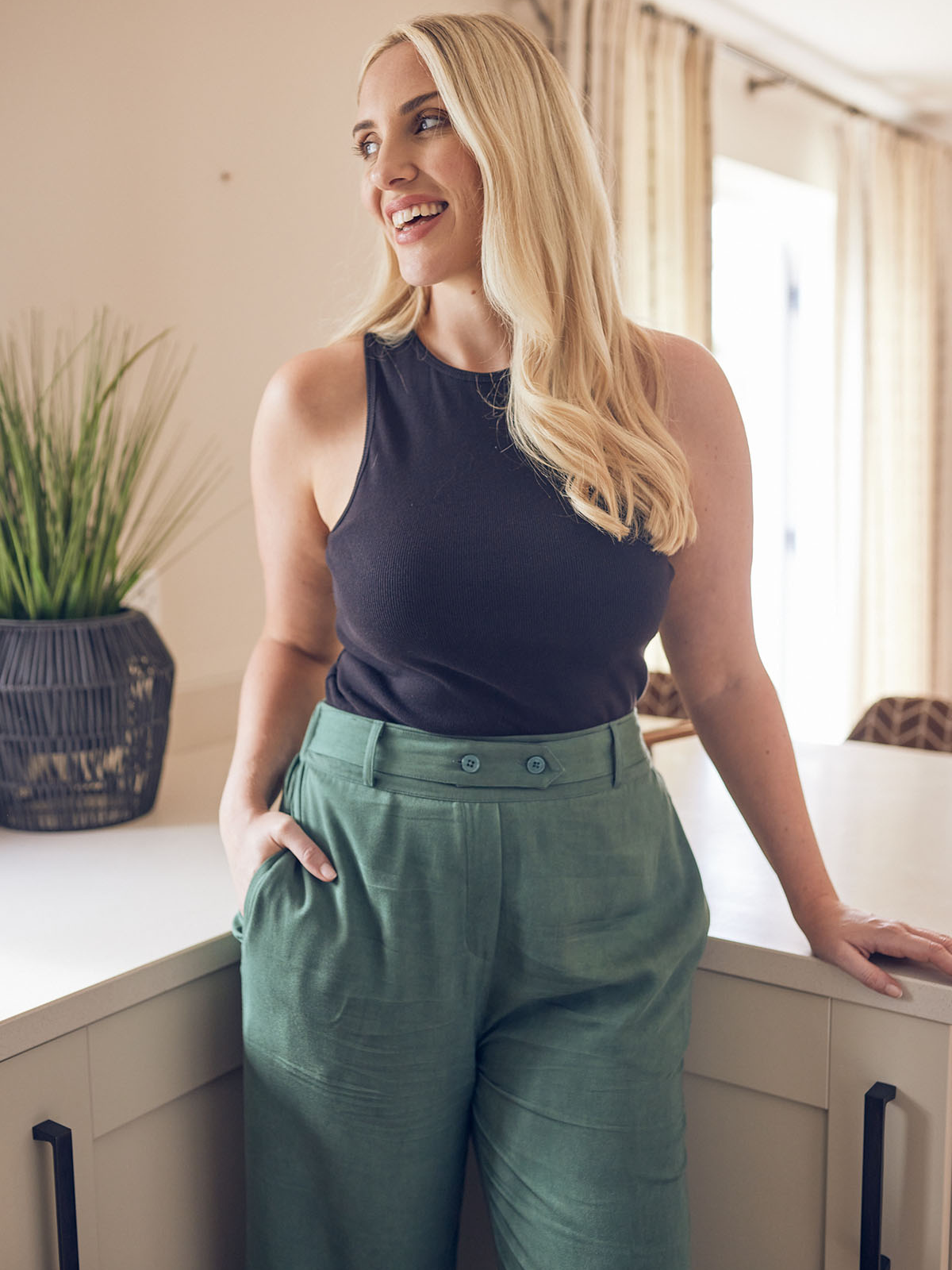 A model is pictured in an open plan kitchen area leaning against a counter top. They are wearing the Abbey rib vest and olive Beth trousers, with one hand in their pocket. 