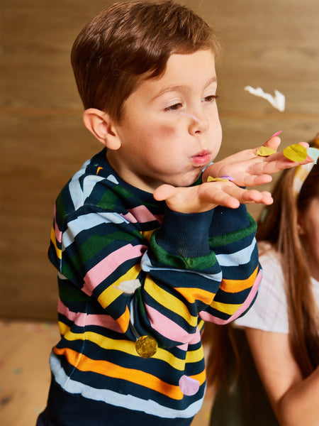 Child wearing navy kids sustainable tracksuit top with a colourful wavy print. Child is blowing confetti and having fun.