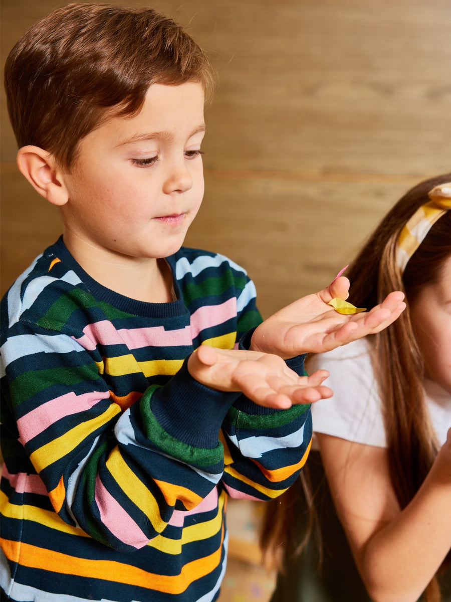 Child wearing sustainable Elias tracksuit for kids, with colourful stripes in pink, yellow, orange, blue and green. Child has outstretched hands and is standing next to another child in sustainable kids clothing from This is Unfolded.