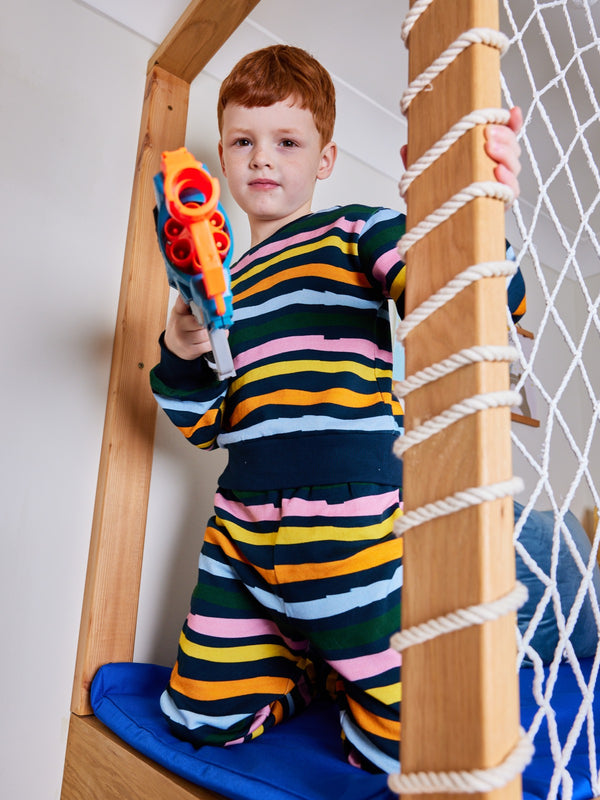 Child wearing the Elias sustainable kids tracksuit set in wavy multicolour stripe, pictured playing with a toy on an indoor climbing frame.
