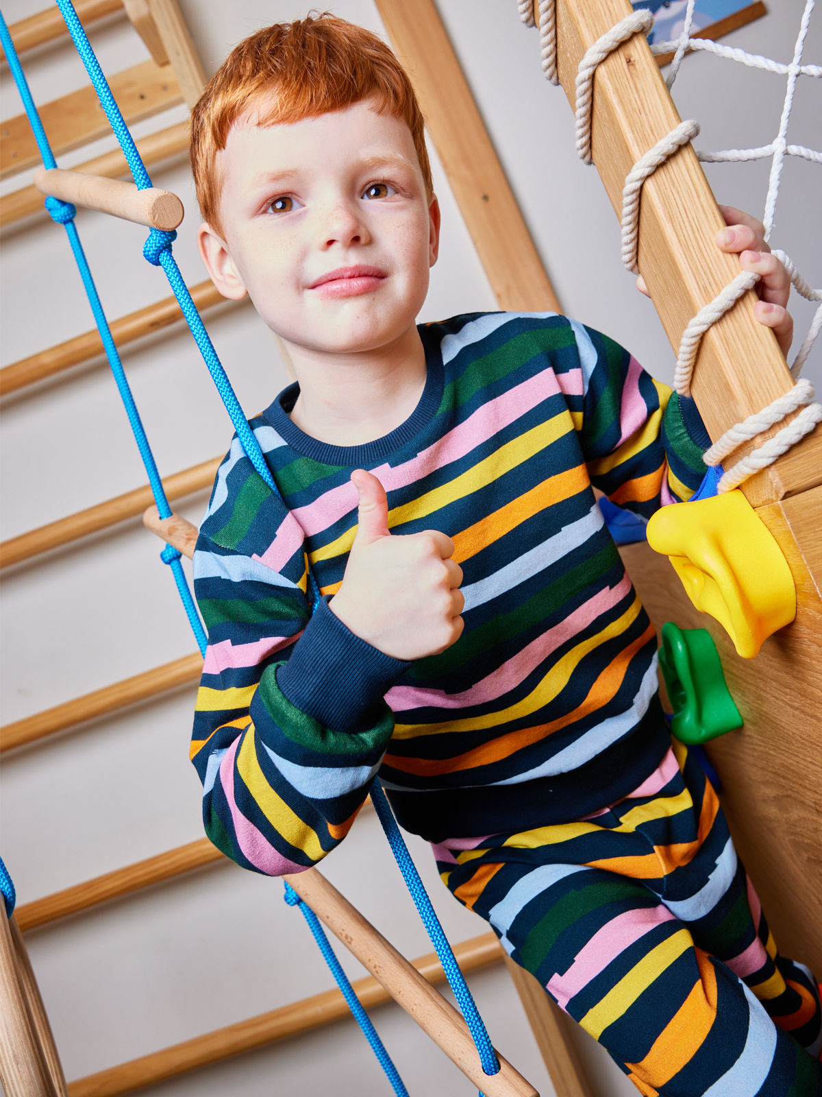 Child wearing the Elias sustainable kids tracksuit set in wavy multicolour stripe, pictured playing on an indoor climbing frame and giving a thumbs up to the camera. 