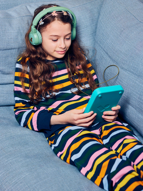 Child wearing the Elias sustainable kids tracksuit set in wavy multicolour stripe, pictured lying on a couch wearing headphones, and looking at an audiobook player