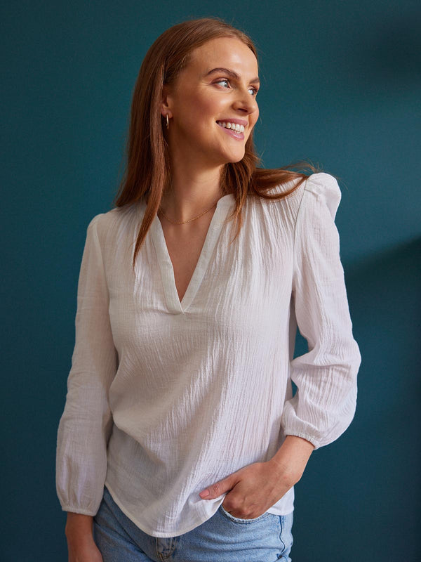 A model wears the white Eve blouse from Unfolded against a backdrop of a dark teal wall. They have one of their hands in their jeans pocket and are smiling to the side. 