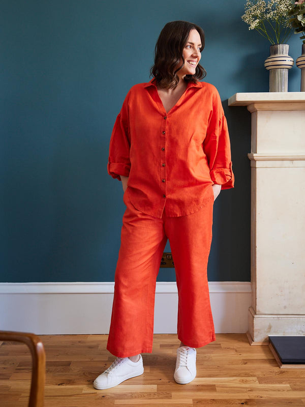 A model wears both the Gigi trousers and the Martha blouse in orange-red as a brightly coordinated set. They are pictured in a living area with dark teal walls and a large fireplace with flowers on the mantlepiece.  