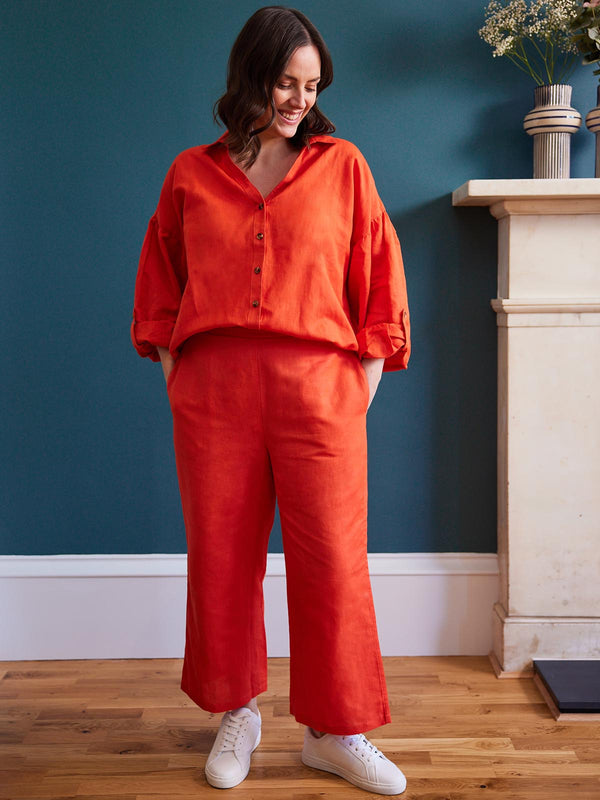 A model wears both the Gigi trousers and the Martha blouse in orange-red as a brightly coordinated set. They are pictured in a living area with dark teal walls and a large fireplace with flowers on the mantlepiece.