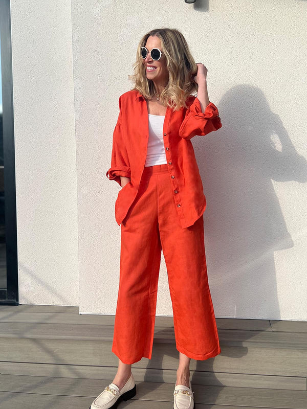 A woman wears the Gigi trousers and the Martha shirt in red, as a coordinating set. She is pictured wearing sunglasses on a sunny deck area outdoors. 