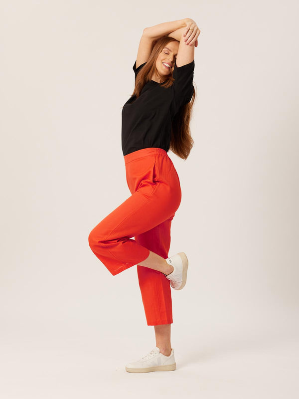 A model wears the Gigi trousers, pictured against a white backdrop and crossing their arms over their head and smiling. 