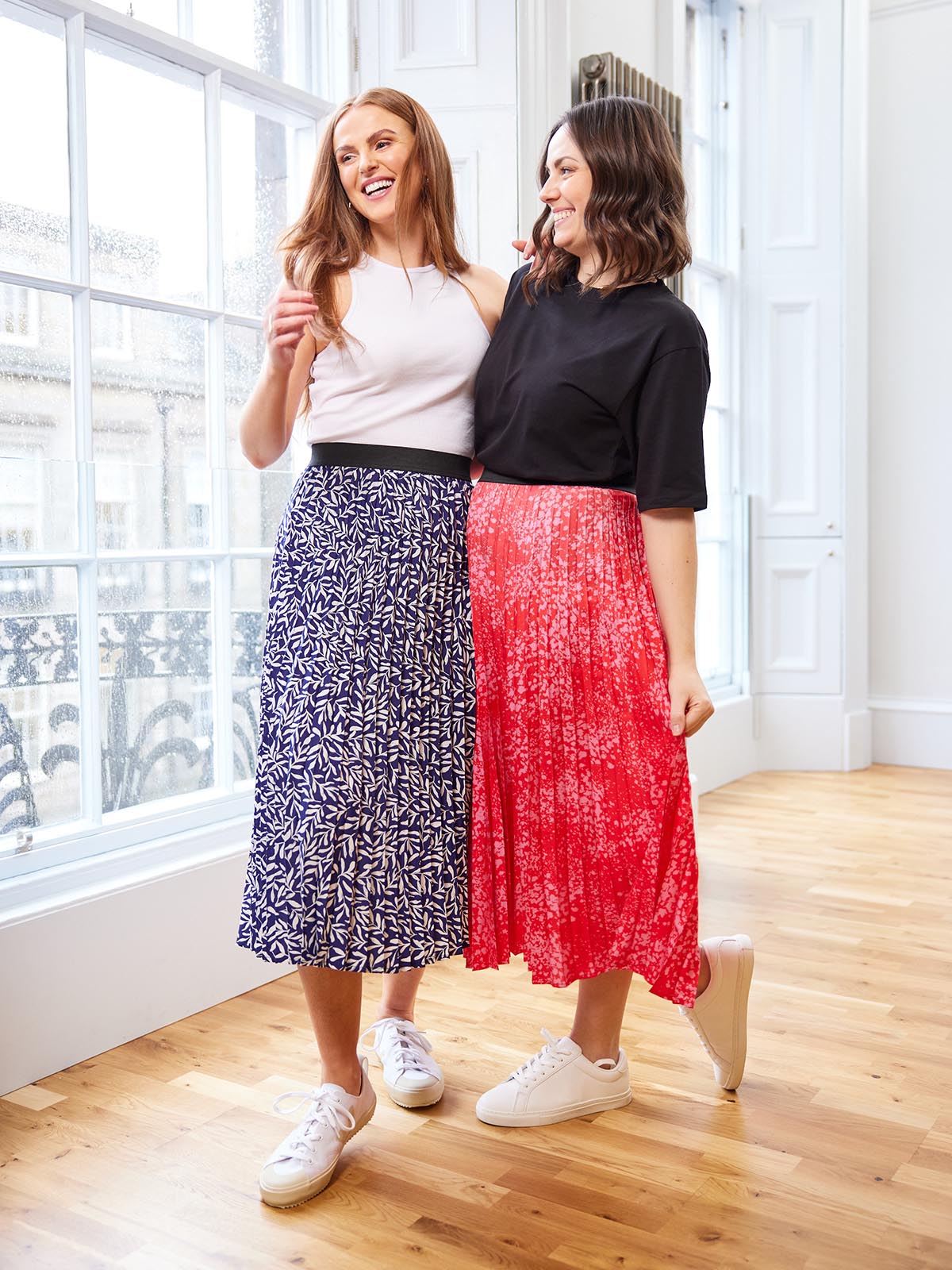 Two models wearing the Gill skirt, one in navy and the other in pink. They are pictured standing close together and one has their arm around the other like good friends. 