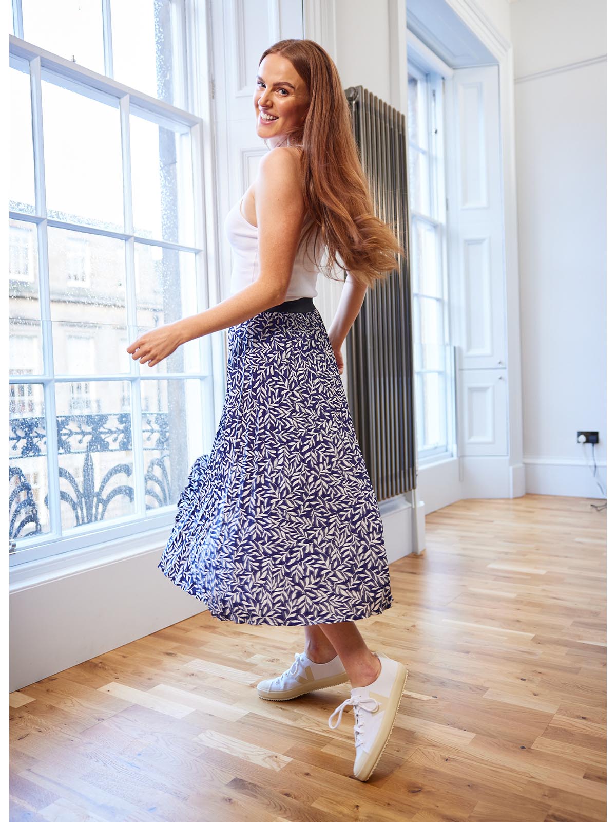 A model wearing the Gill sustainable skirt in navy and a white vest top, pictured in a well lit room in front of a large window and smiling. 