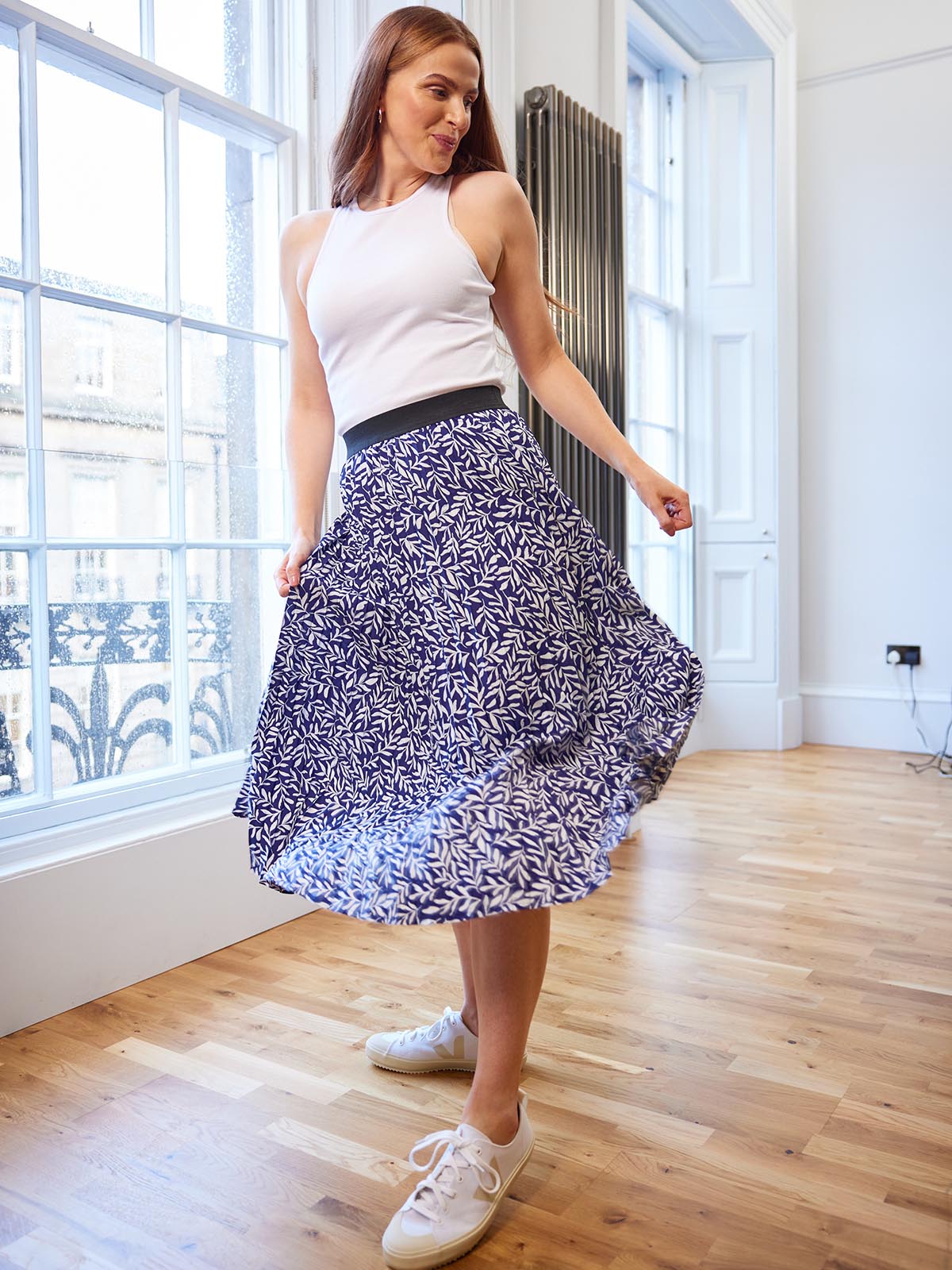 A model wearing the Gill sustainable skirt in navy and a white vest top, pictured in a well lit room in front of a large window and swishing the skirt.
