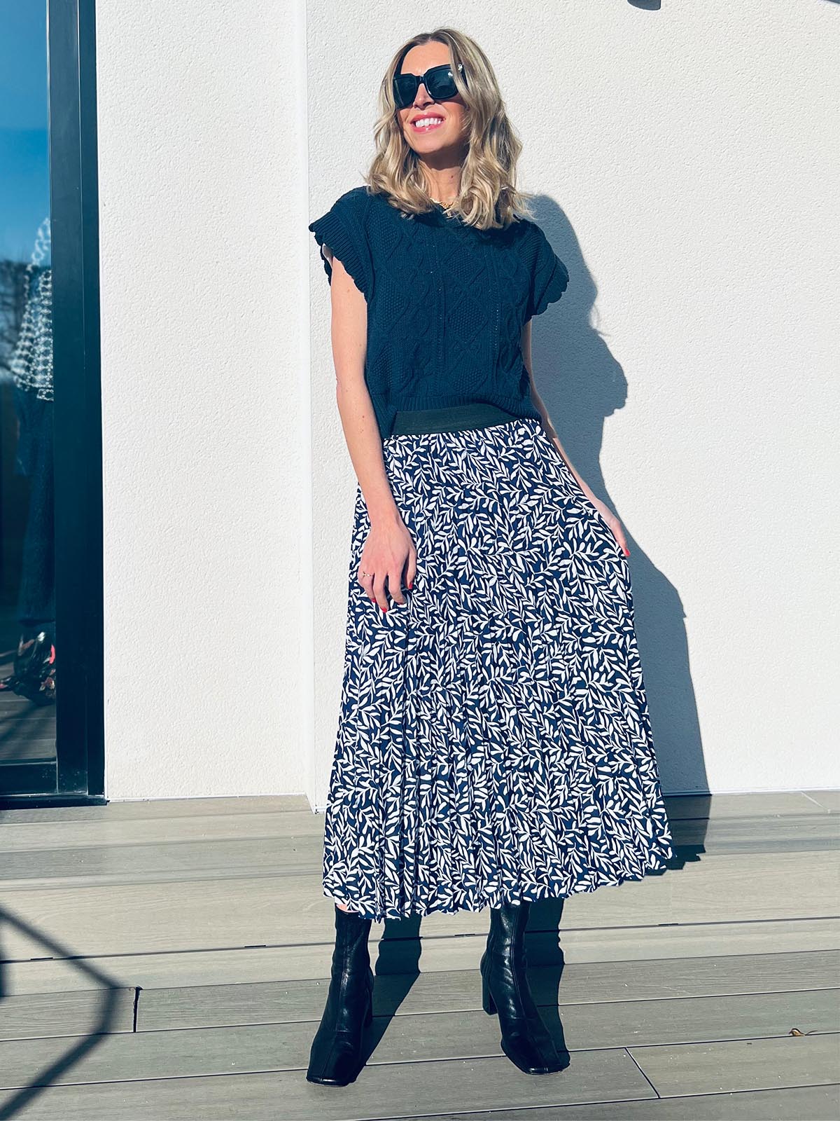 A model wearing the pleated Gill skirt in navy with a short sleeve t-shirt, pictured wearing sunglasses on a balcony on a sunny day.