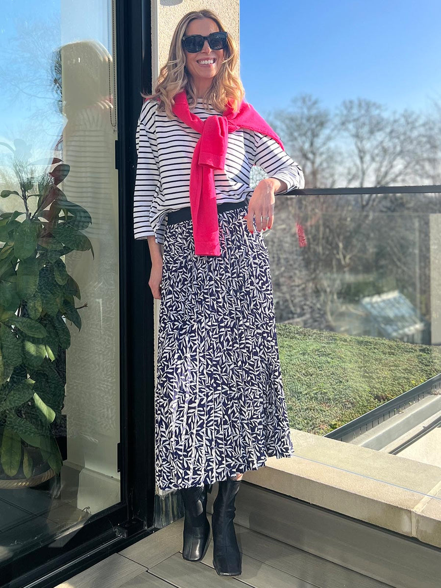 A model wearing the pleated Gill skirt in navy with a striped t-shirt, pictured wearing sunglasses on a balcony on a sunny day.