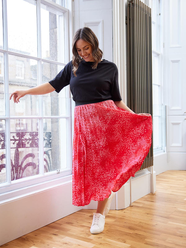 A model pictured in front of a large window in a well-lit room, wearing the Gill skirt in pink and red and smiling at the ground. 