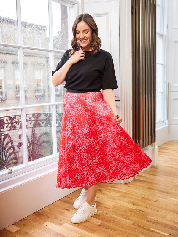 A model pictured in front of a large window in a well-lit room, wearing the Gill skirt in pink and red and smiling at the ground.