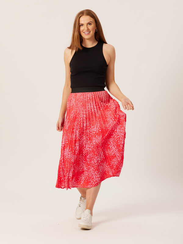 A model wearing the Gill skirt in red and pink smudge print, pictured walking towards the camera and causing movement in the skirt pleats. 