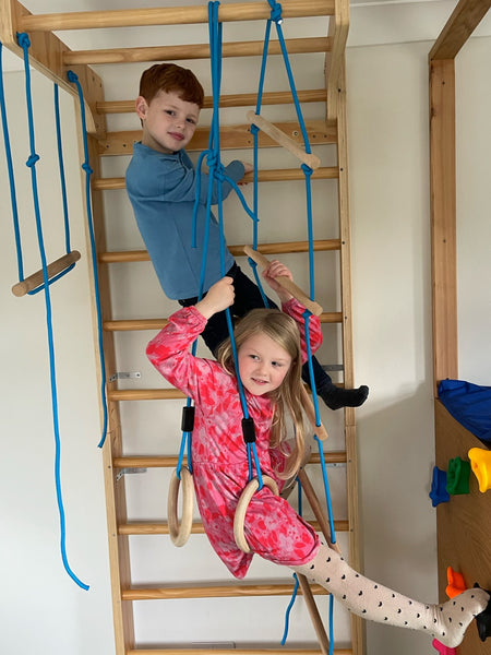 Two children, one wearing the sustainable Harris kids button down t-shirt and one wearing the Izzy eco-friendly kids smock dress, pictured playing on an indoor climbing frame together. 