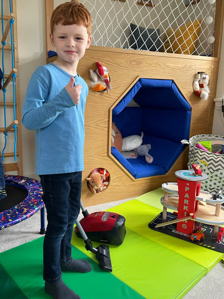 A child wearing the Harris sustainable kids pique t-shirt in blue, pictured in a playroom playing with a toy vacuum cleaner and holding their thumb up whilst smiling at the camera.