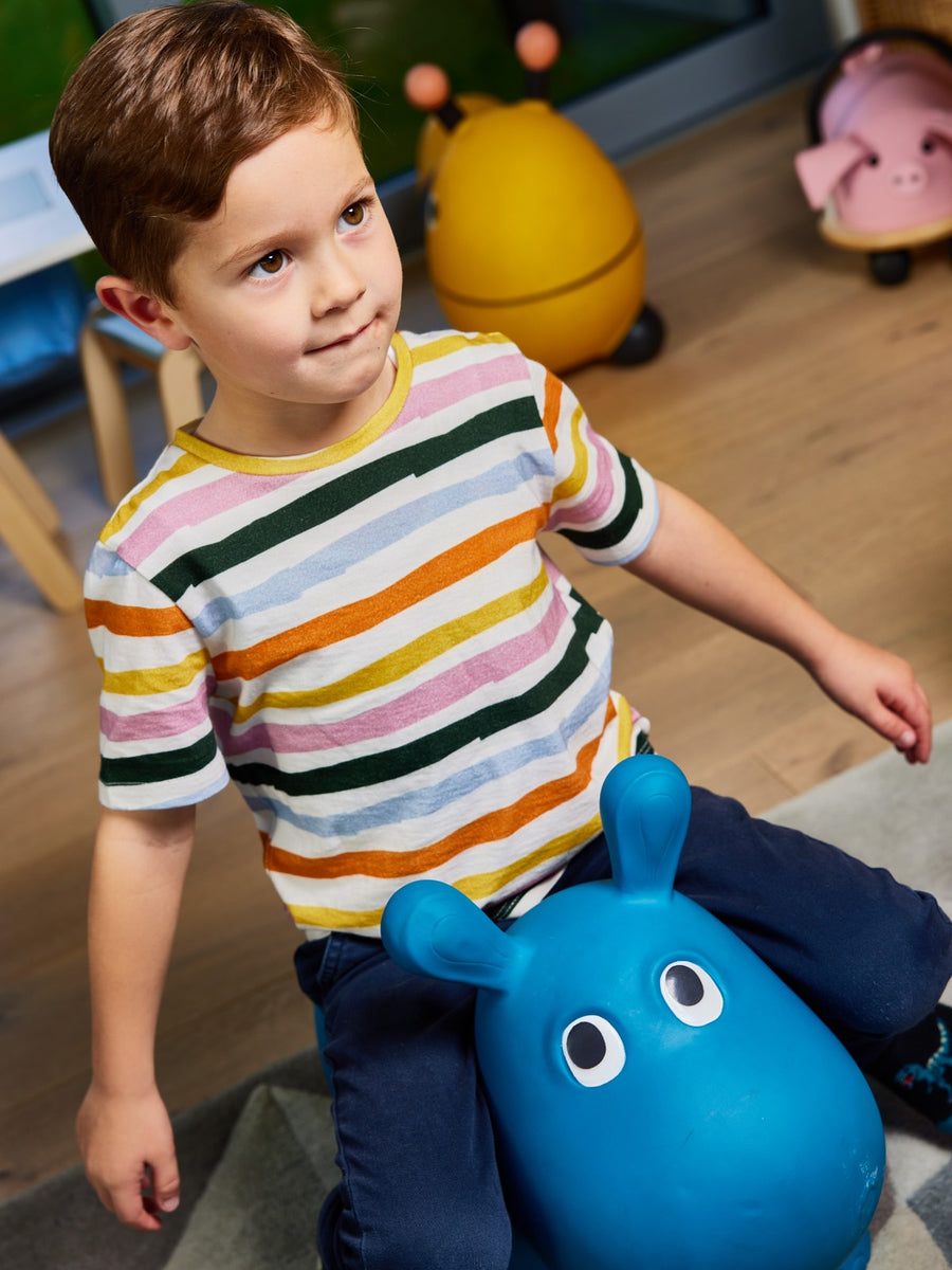 A child wearing the Hedy sustainable unisex kids t-shirt in multicolour stripe, pictured in a playroom sitting on a toy hippo. 