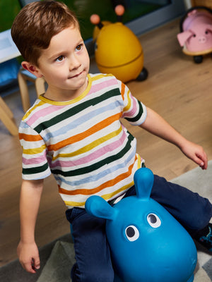 A child wearing the Hedy sustainable unisex kids t-shirt in multicolour stripe, pictured in a playroom sitting on a toy hippo. 