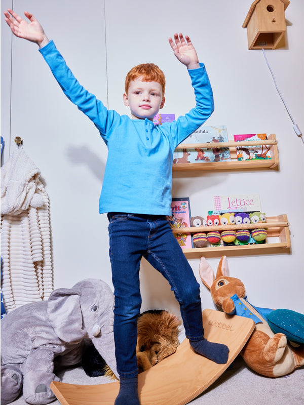 A child wearing the Harris sustainable kids pique t-shirt in blue, pictured standing on a wooden balance board and waving their arms. 