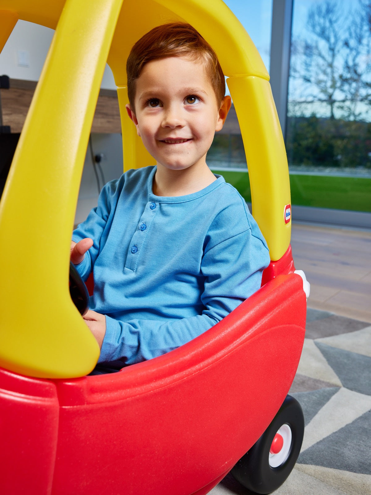 A child pictured driving a red and yellow toy car, wearing the Harris sustainable kids pique t-shirt in blue from This is Unfolded. 