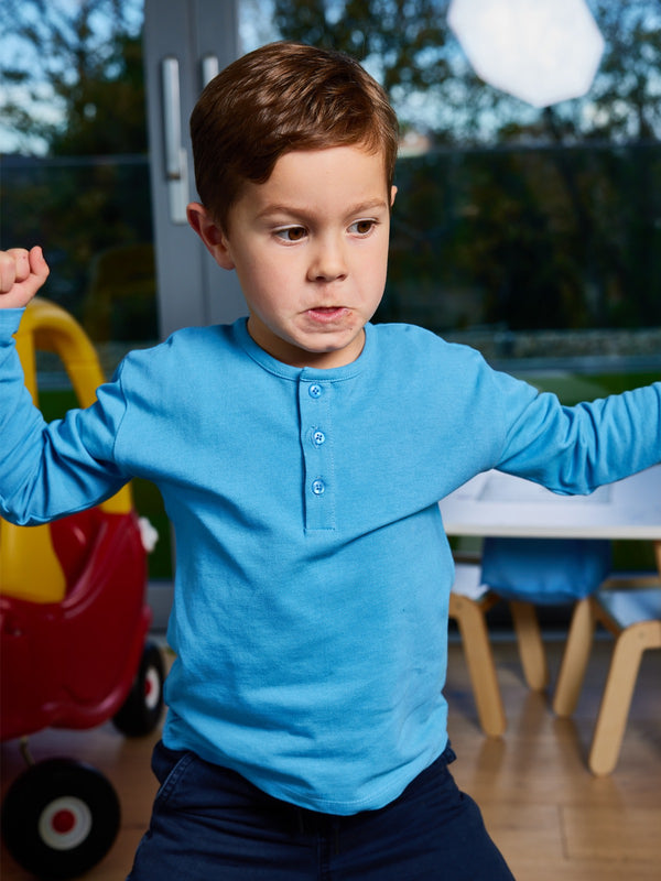 A child wearing the Harris sustainable kids pique t-shirt in blue, pictured in a playroom with their arms raised.
