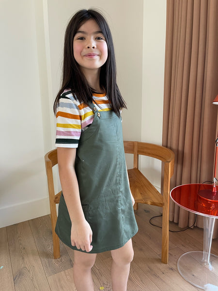 A child wearing the Hedy sustainable unisex kids t-shirt in multicolour stripe underneath the eco-friendly Lilah kids pinafore, pictured looking into the camera and smiling. 