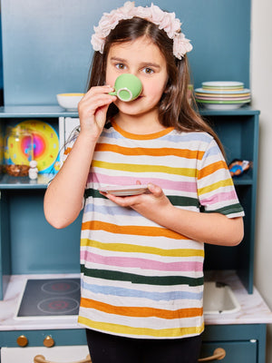 A child wearing the Hedy sustainable unisex kids t-shirt in multicolour stripe, pictured sipping from a children's tea set and looking  at the camera.