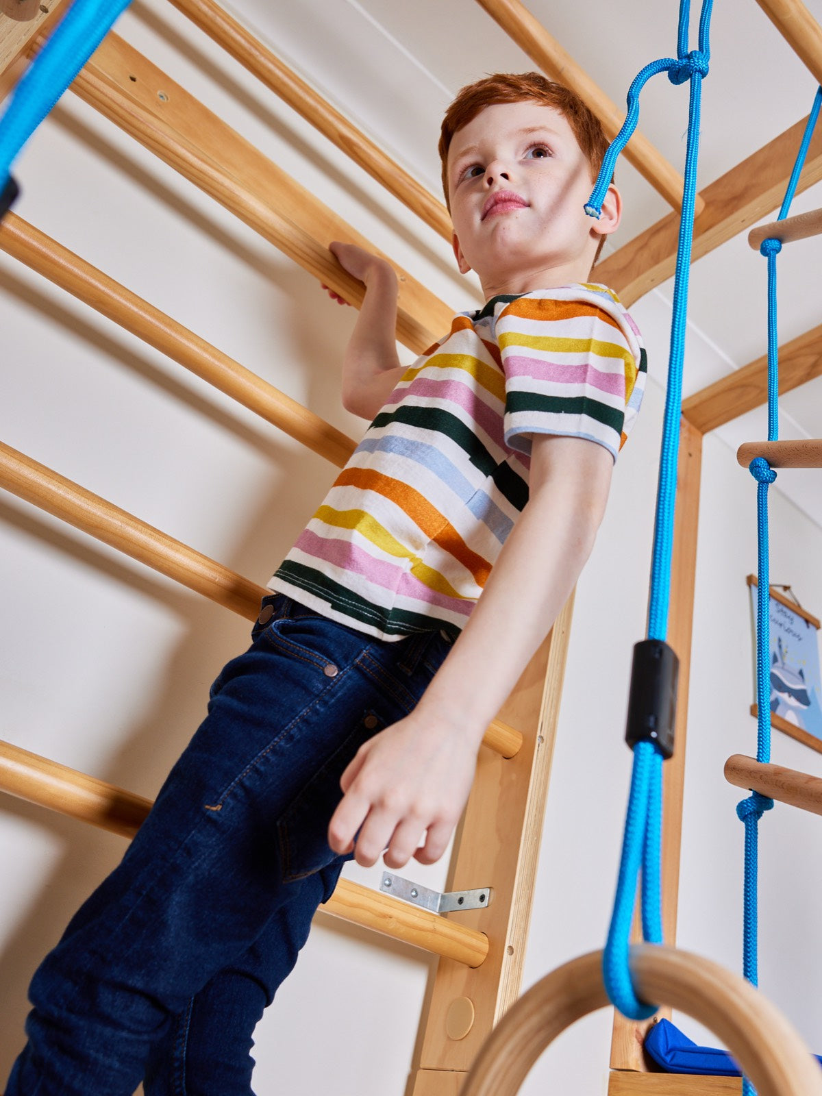 A child wearing the Hedy sustainable unisex kids t-shirt in multicolour stripe, pictured climbing on an indoor climbing frame.