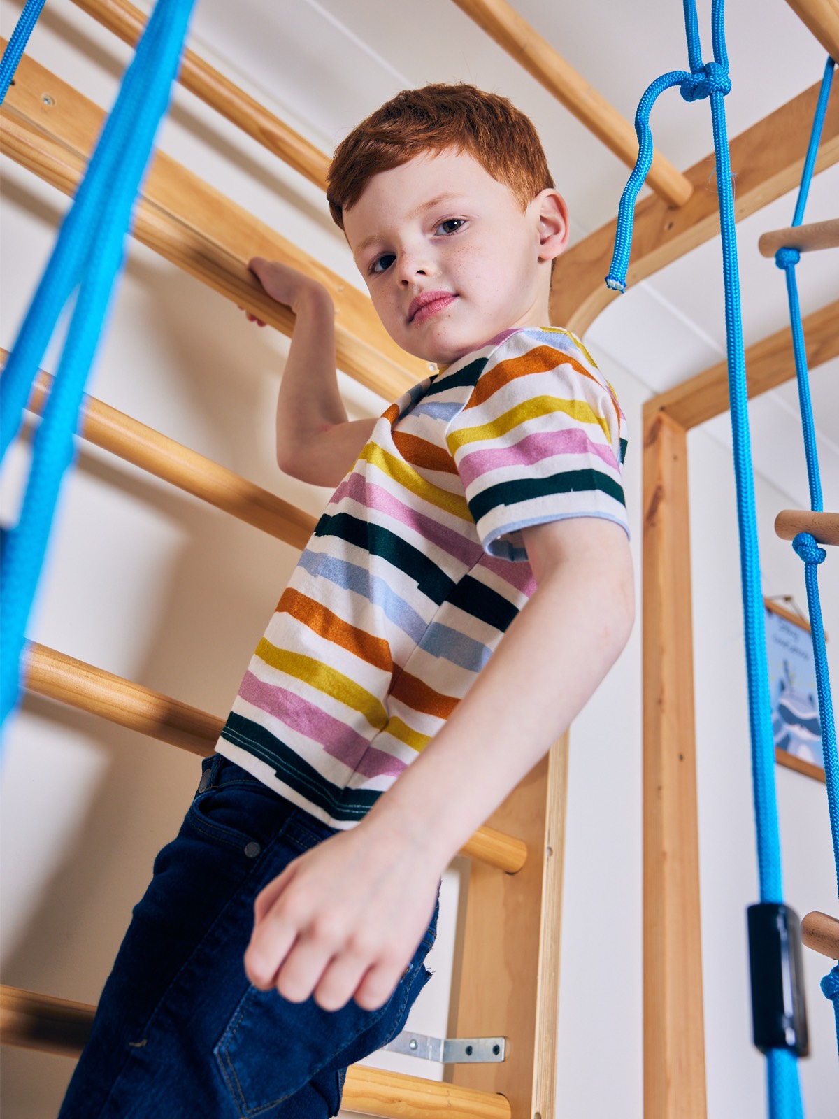 A child wearing the Hedy sustainable unisex kids t-shirt in multicolour stripe, pictured climbing on an indoor climbing frame and looking into the camera.