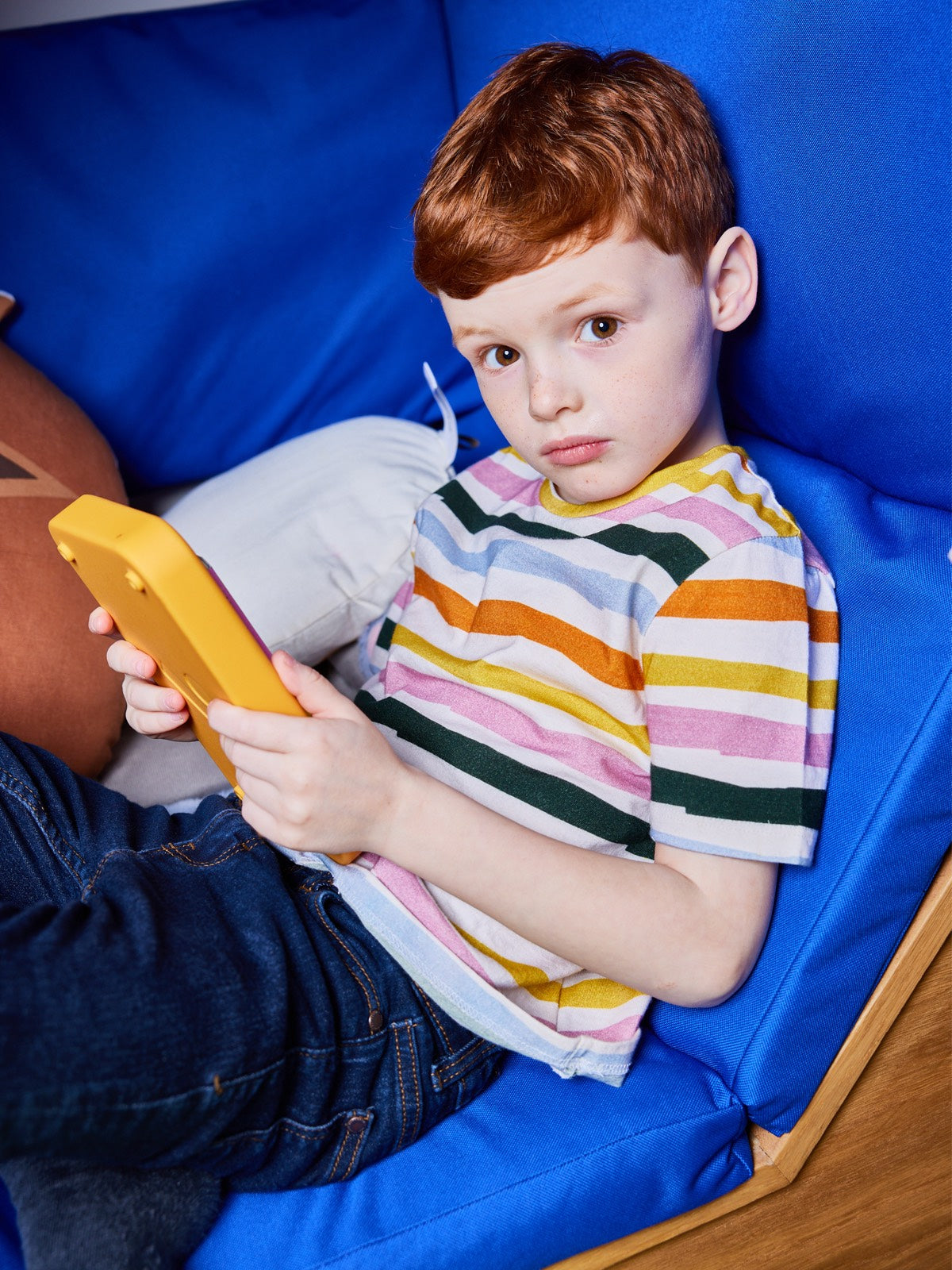 A child wearing the Hedy sustainable unisex kids t-shirt in multicolour stripe, pictured lying on a seating area and holding a tablet whilst looking into the camera.