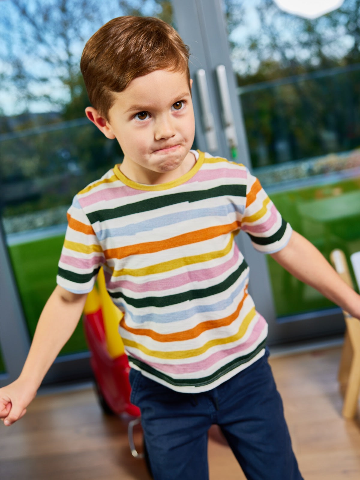 A child wearing the Hedy sustainable unisex kids t-shirt in multicolour stripe, pictured in a playroom with their arms raised at their sides. 