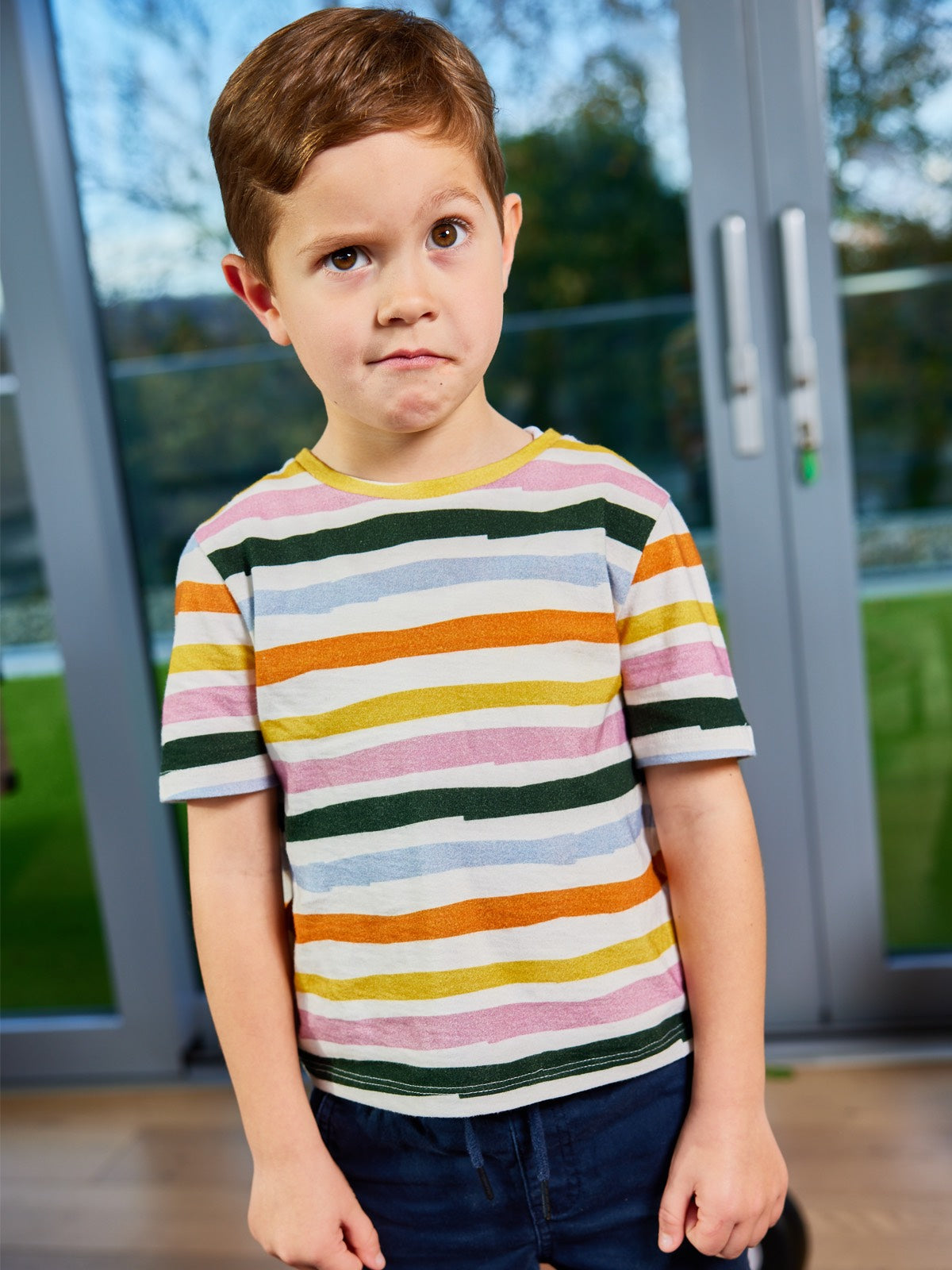 A child wearing the Hedy sustainable unisex kids t-shirt in multicolour stripe, pictured pulling a cheeky face. 