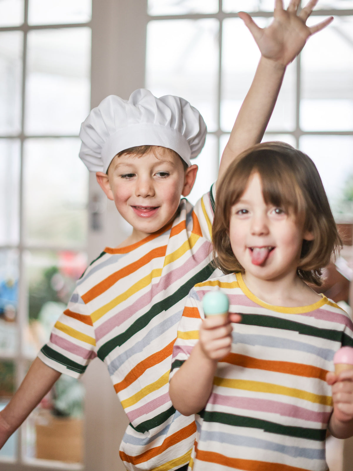 Two children playing ice cream shop while wearing unisex sustainable T-shirts from This is Unfolded. T-shirts are white with colourful wavy stripe patterns.