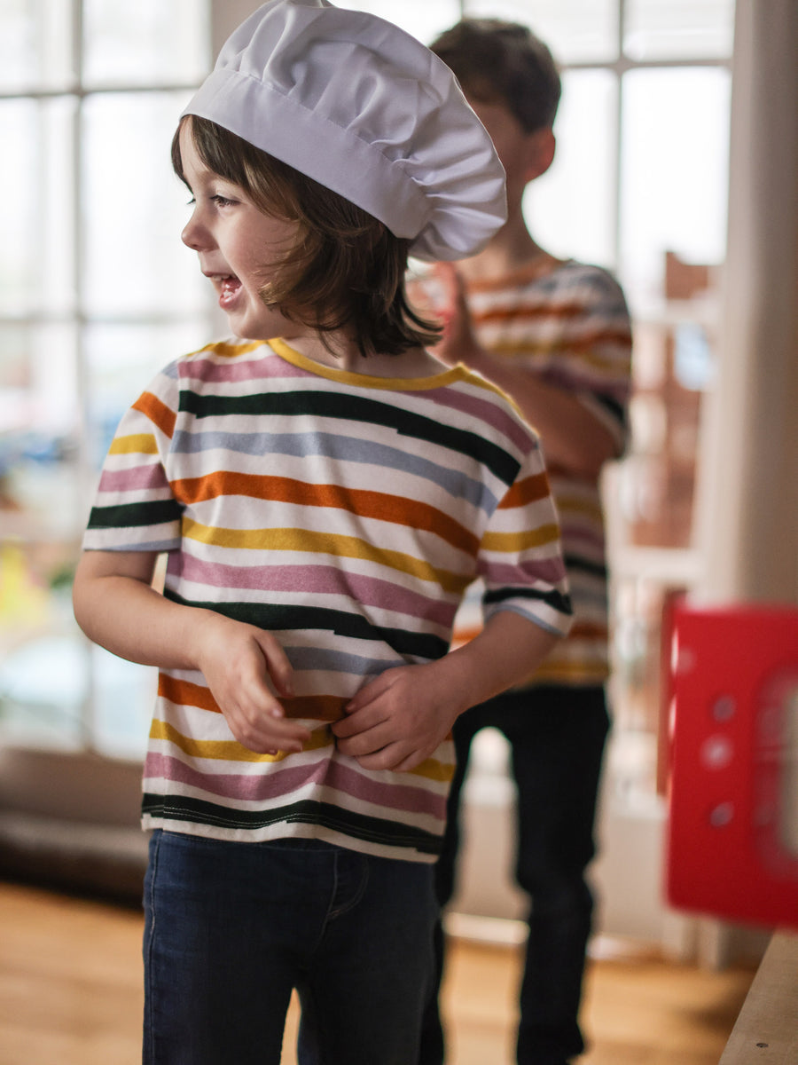 Child wearing sustainable stripy kids t-shirt from This is Unfolded and a chefs hat while playing. The T-shirt has colourful stripes and another child can be seen in the same eco-friendly T-shirt in the background.