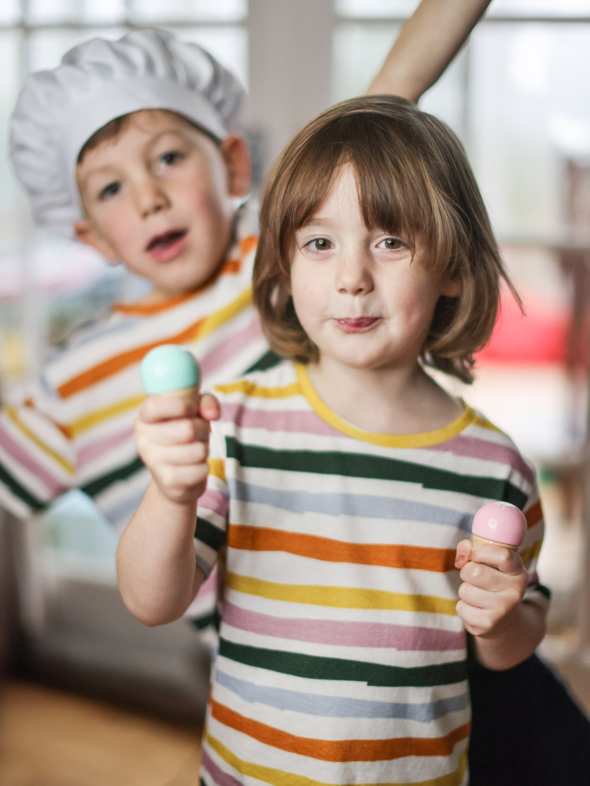 Two playful children wearing the sustainable kids T-shirt Hedy from This is Unfolded. They're playing and goofing around with toy ice creams and chefs hat. The T-shirts are colourful stripy unisex tees for kids.