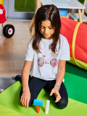 A child wearing the Ivy eco-friendly kids t-shirt with a multicolour bicycle print, pictured on a floormat playing with colourful wooden toy blocks. 