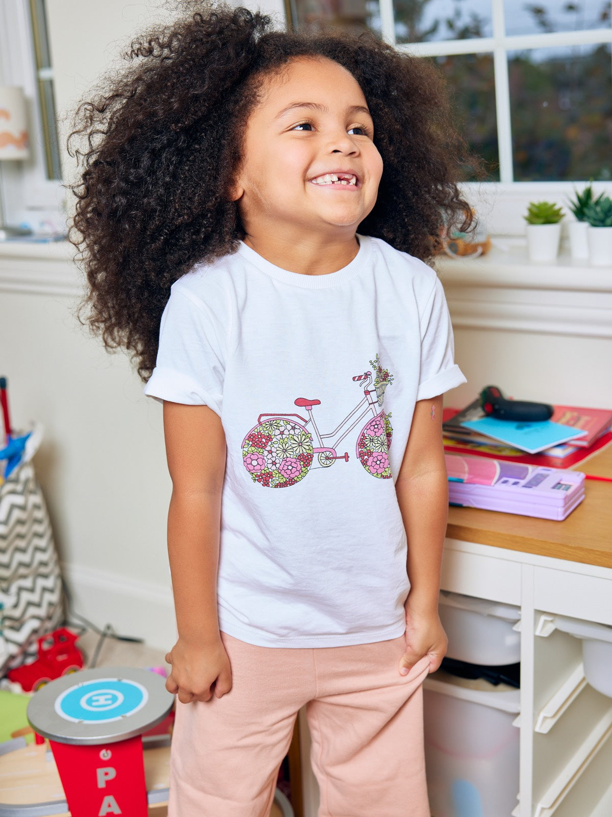 A child pictured in a playroom in front of a desk and smiling, wearing the Ivy eco-friendly kids t-shirt with a multicolour bicycle print from This is Unfolded. 