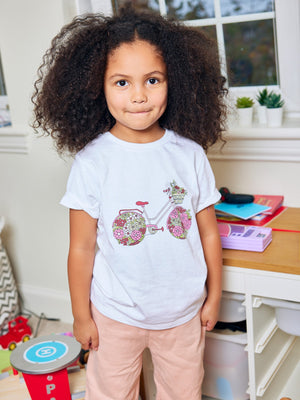 A child wearing the Ivy eco-friendly kids t-shirt with a multicolour bicycle print, pictured in a playroom and looking into the camera with a smile. 