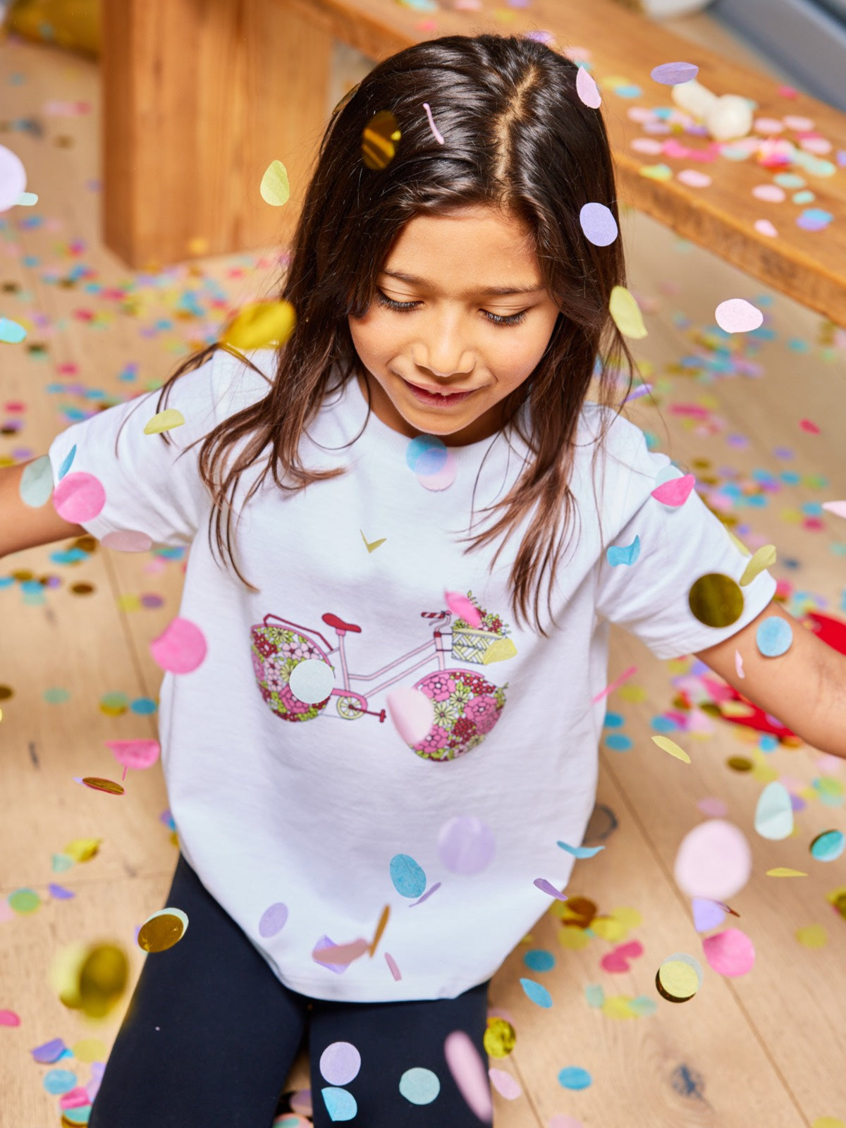A child wearing the Ivy eco-friendly kids t-shirt with a multicolour bicycle print, pictured with falling confetti in a party atmosphere. 