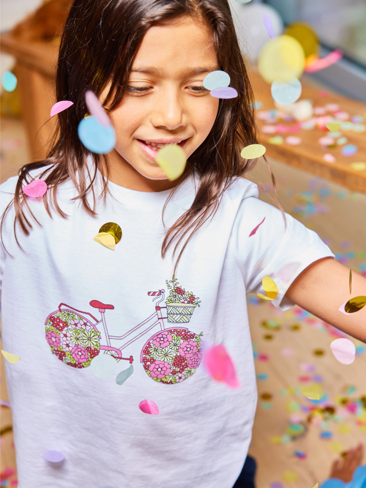 A child pictured with falling confetti wearing the Ivy sustainable kids t-shirt from This is Unfolded, with a bicycle print in pinks and green.