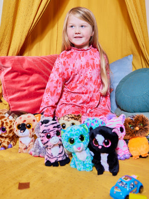 A child wearing the Izzy eco-friendly kids smock dress in pink, pictured in a bright playroom surrounded by cuddly toys and cushions.