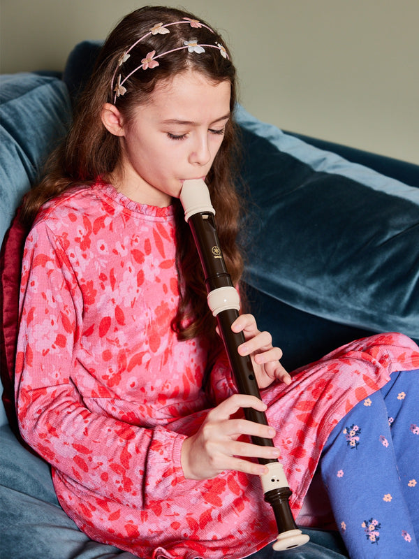 A child wearing the Izzy eco-friendly kids smock dress in pink, pictured sitting on a couch and playing a recorder. 