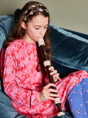 A child wearing the Izzy eco-friendly kids smock dress in pink, pictured sitting on a couch and playing a recorder. 