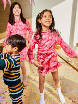 Three children, two wearing the Izzy eco-friendly kids smock dress in pink and one wearing the Elias sustainable tracksuit set in wavy multicolour stripe, pictured together in a room with confetti on the floor. 