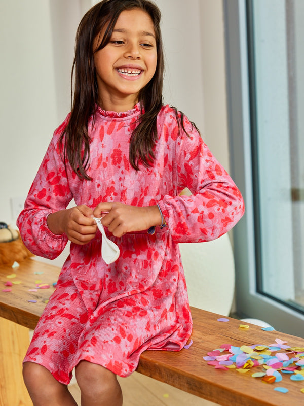 A child wearing the Izzy sustainable kids smock dress in pink, pictured playing with a blow-up balloon and surrounded by confetti. 