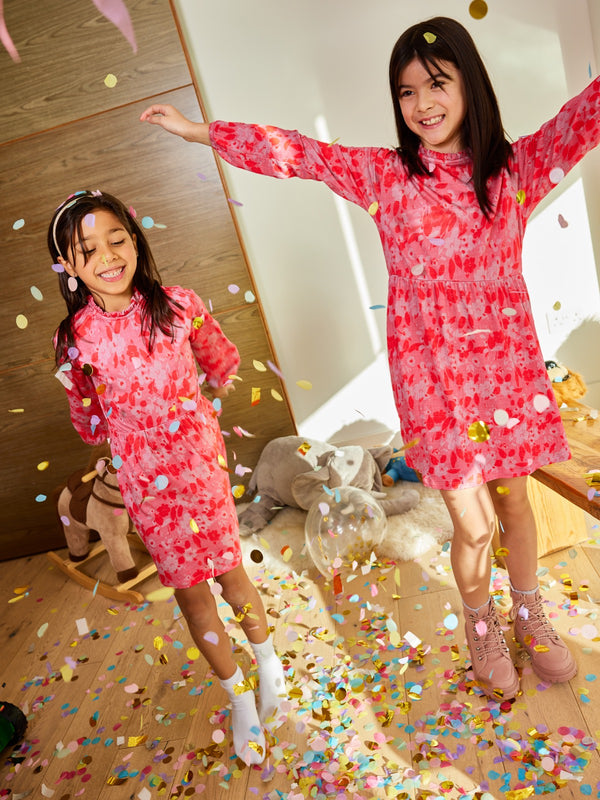 Two children wearing the Izzy sustainable kids smock dress in pink, pictured with falling confetti and smiling. 