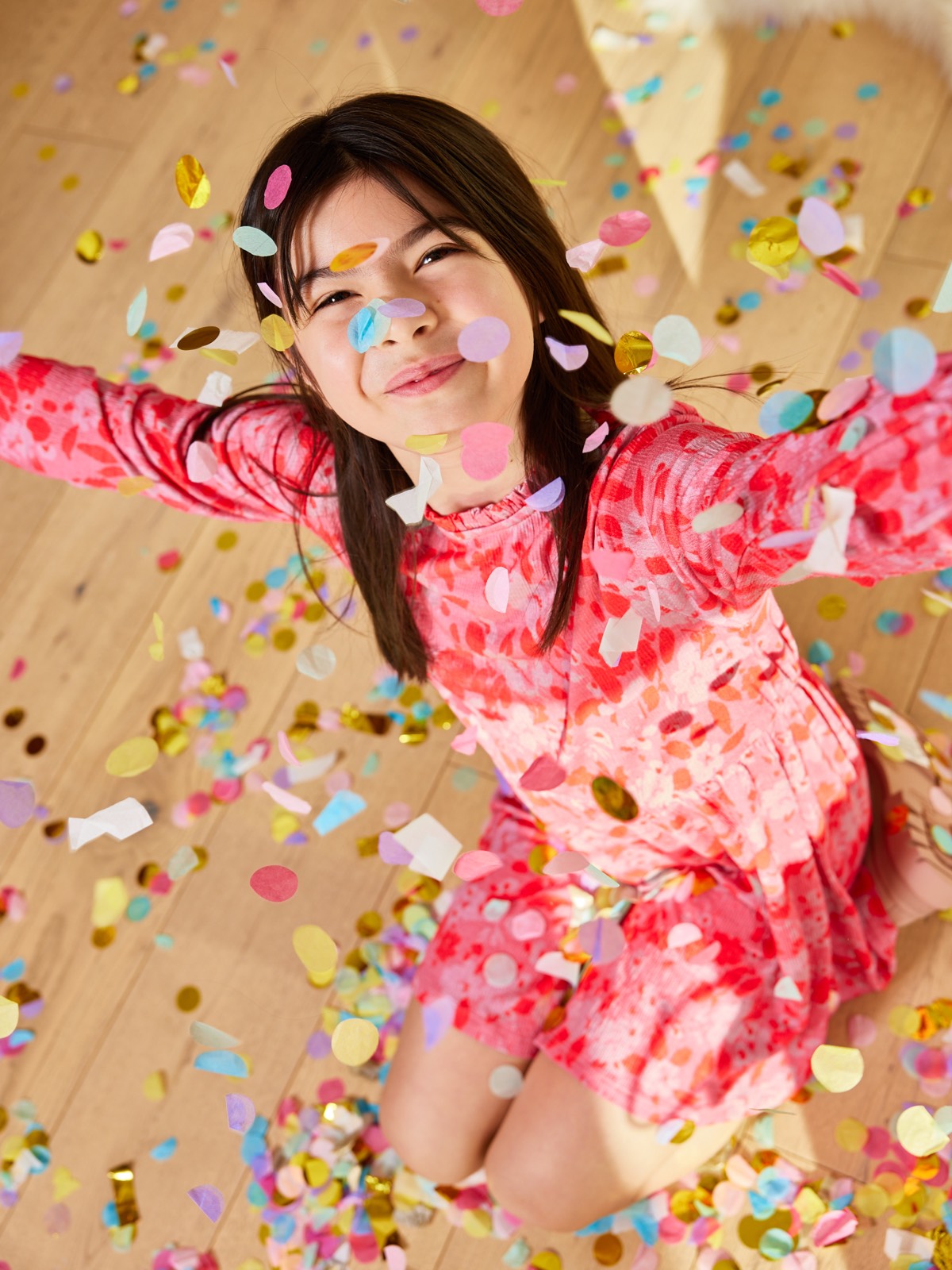 A child wearing the Izzy eco-friendly kids smock dress in pink, pictured with falling confetti and smiling at the camera.